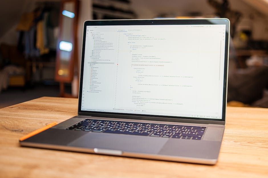 A man with glasses and a beard is seated at a wooden desk working on a computer with dual monitors, both displaying lines of code and software interfaces. The workspace is organized, with a keyboard, 